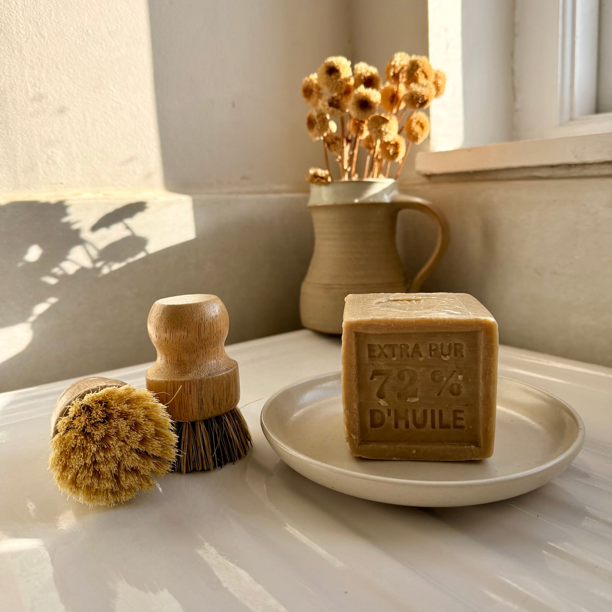 A rectangular bar of natural soap labeled 'EXTRA GOUR 72% D'HUILE' sits on a small white dish, next to a wooden-handled brush and a dried flower arrangement in a ceramic pot on a windowsill.