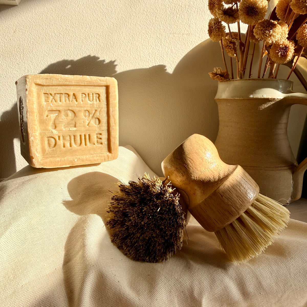 A natural skincare set featuring a square bar of olive oil soap embossed with 'EXTRA PUR 72% D'HUILE', a wooden-handled brush with natural bristles, and a dark brown scrubber, styled on a cream fabric surface next to a ceramic vase with dried flowers.