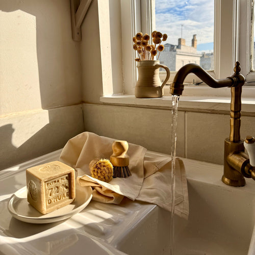 A rustic kitchen sink scene with running water from a brass faucet, a beige soap dish holding a bar of soap, a folded beige towel, and a natural bristle brush. A ceramic mug with brushes sits on the windowsill beside a white-framed window showing blue sky and rooftops.