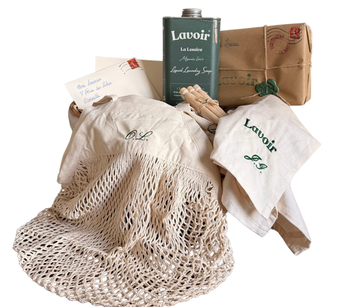 A vintage-style laundry care set featuring a cream-colored mesh laundry bag, a matching embroidered cotton cap and bandana, a green vintage canister of Lavoir liquid laundry soap, a brown envelope with a postmark, and a wooden clothes hanger, all arranged on a white background.